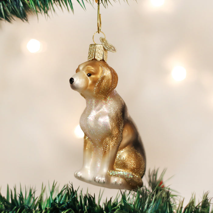 A Christmas tree ornament shaped like a sitting dog, decorated with gold and white glitter, hangs from a branch with blurred lights in the background.