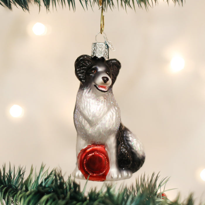 A Christmas tree ornament shaped like a black and white dog with a red ribbon sits on a green branch, with blurry white lights in the background.