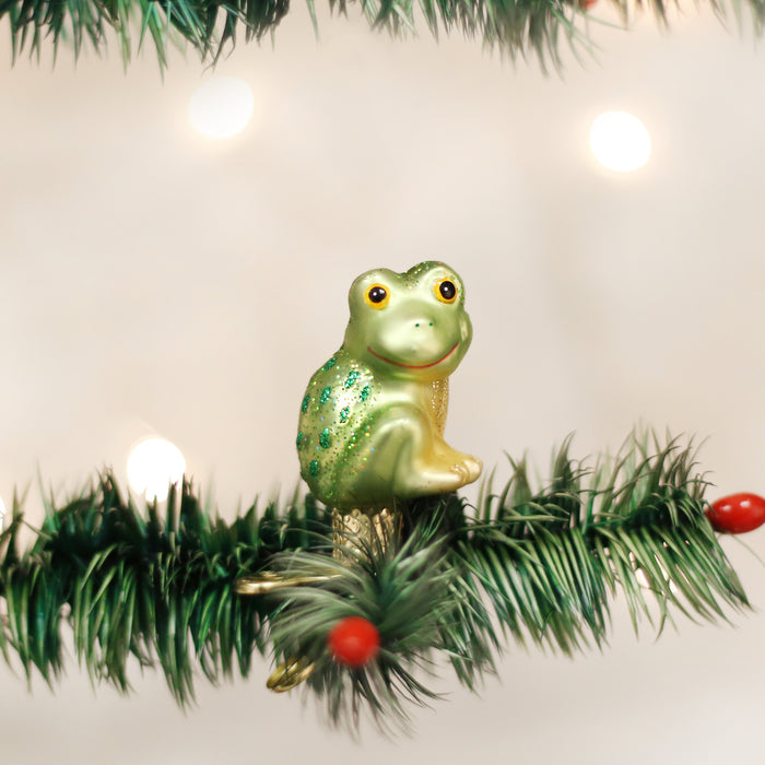 A green frog-shaped ornament with glitter details sits on a pine branch, decorated with small red berries, against a blurred background of white lights.