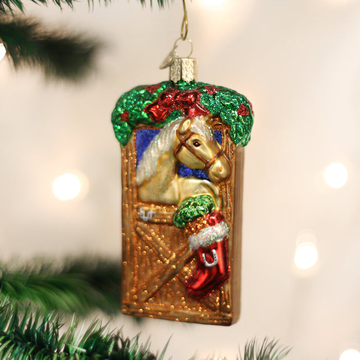 A Christmas tree ornament shaped like a stable door, featuring a tan horse peeking out, decorated with greenery, red bows, and a red stocking. Tree branches and white lights are blurred in the background.