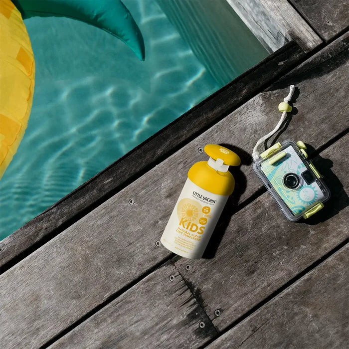 A bottle of sunscreen and a waterproof camera resting on a wooden deck next to a swimming pool.