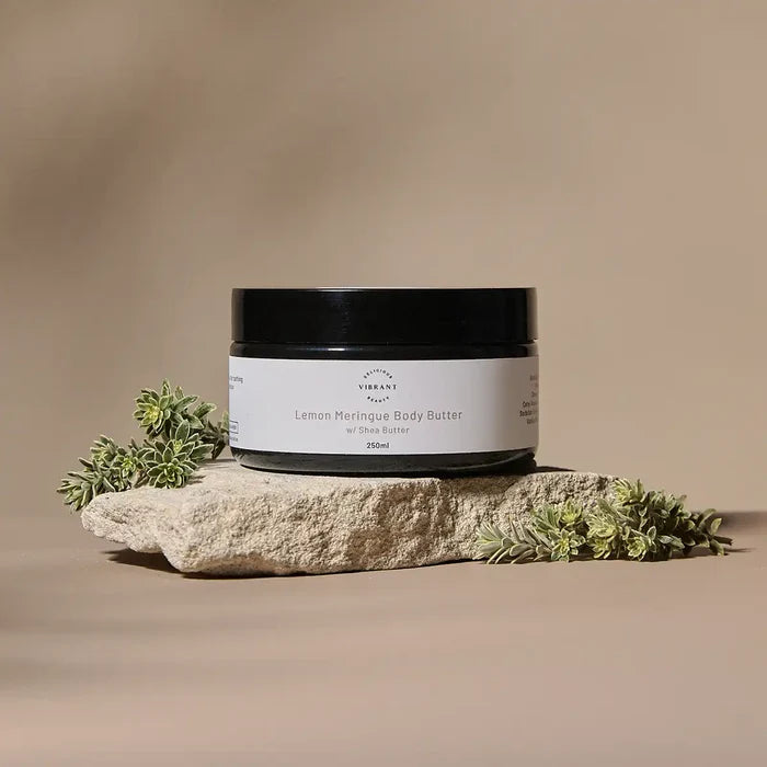 A jar of Lemon Meringue Body Butter sits on a textured stone slab with small green leaves, set against a warm beige background.