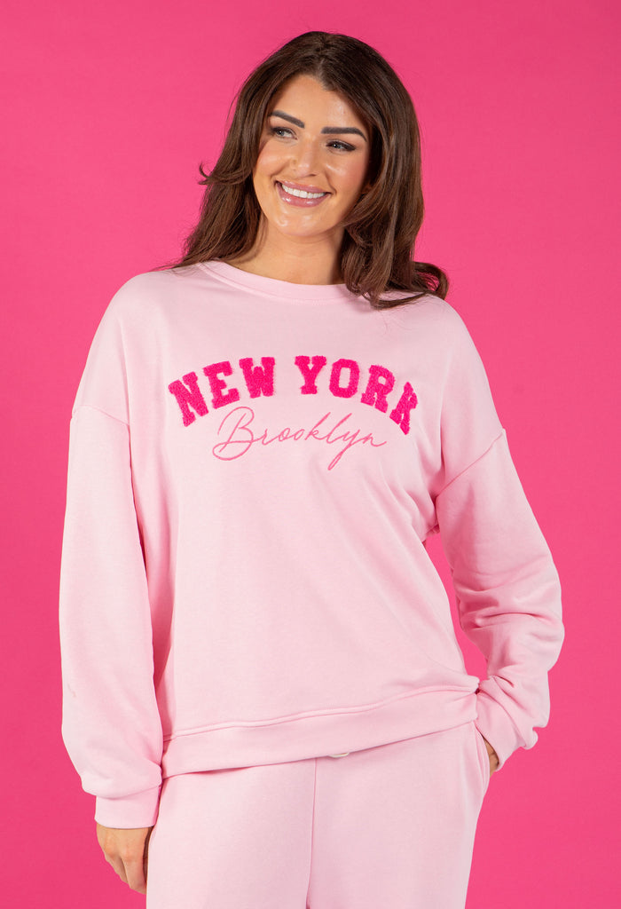 Woman wearing a pink 'New York Brooklyn' sweatshirt against a pink background