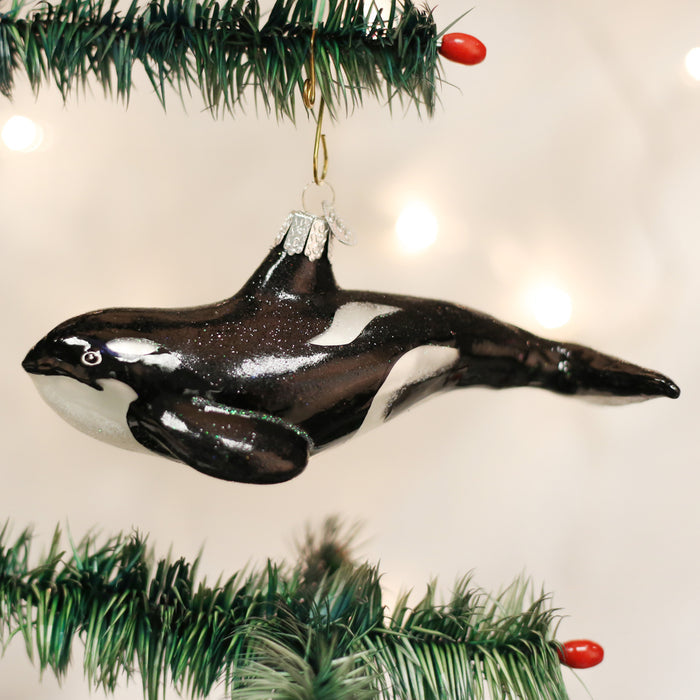 A shiny black and white orca whale ornament hangs from a Christmas tree branch decorated with green needles and small red berries, with white lights glowing softly in the background.