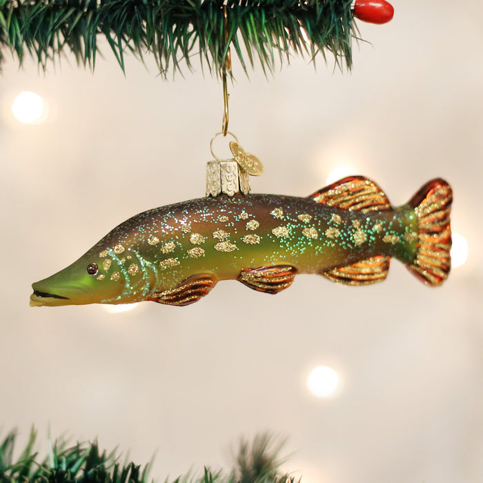 A Christmas tree ornament shaped like a green and brown fish with glittery details hangs from a branch with pine needles and red berries visible.
