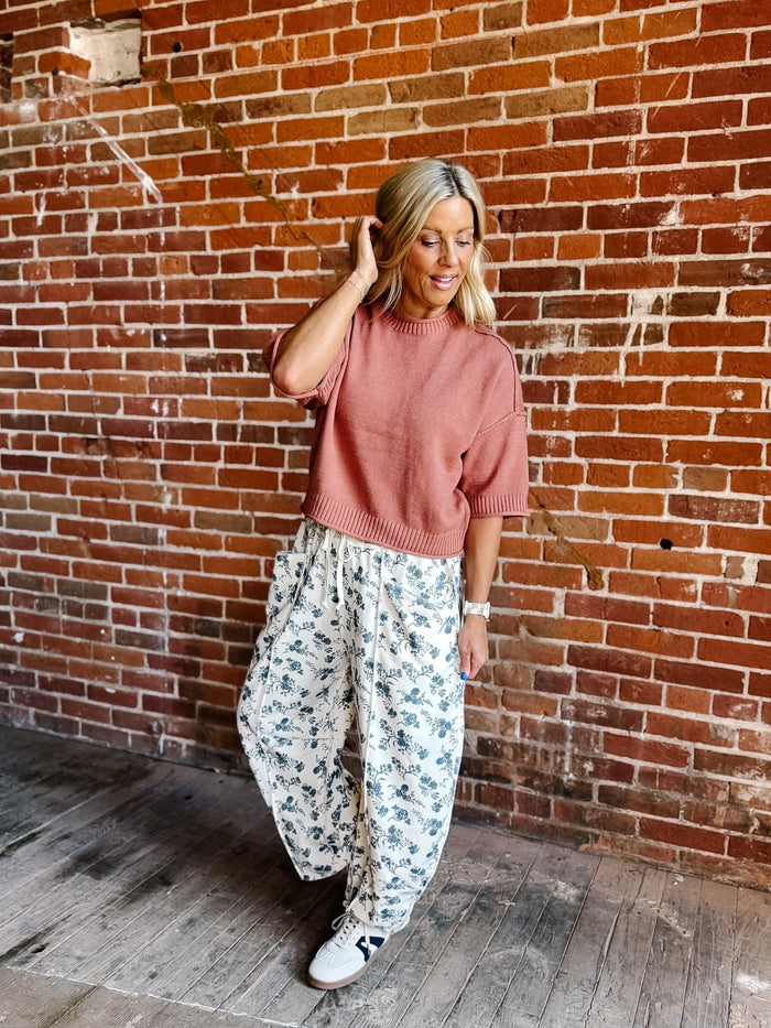 A woman posing in front of a red brick wall, wearing a dusty rose short-sleeve knit sweater and white wide-leg pants with a blue floral pattern, paired with white sneakers.