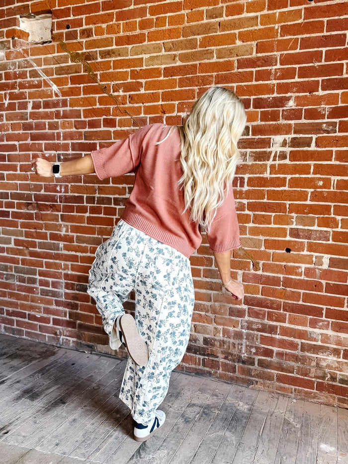 A woman with long blonde hair is shown from behind, wearing a dusty rose short-sleeve knit top and white wide-leg pants with a blue floral pattern, standing against a red-brown brick wall.
