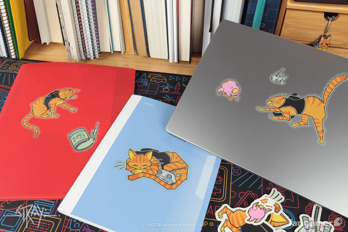 Stray stickers photographed on two types of folder and a Macbook, showing their size. Everything's sitting atop a Walled City 99 desk mat.