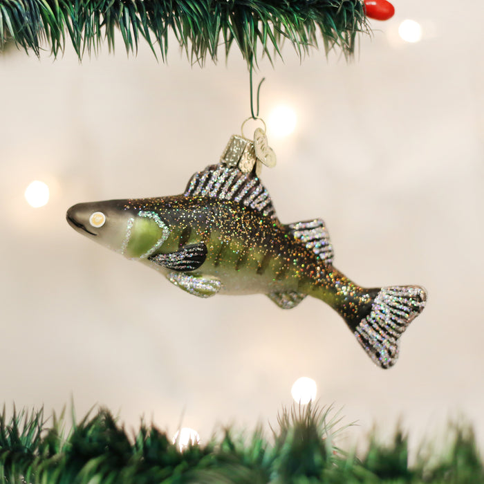 A glittery fish-shaped Christmas ornament hangs from a green artificial tree branch, with blurred white holiday lights in the background.
